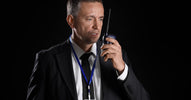 An older male FBI agent is wearing a black suit and tie with a blue FBI badge while speaking into a two-way radio.