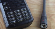 A black two-way radio sits on a light wood table beside its detached radio antenna, with a focus on the keypad and connector.