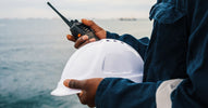 A person in blue coveralls stands on a ship, holding a white hard hat in one hand and a two-way radio in the other.