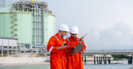 Two plant workers in orange jumpsuits and hard hats review a clipboard and hold two-way radios outside a power plant.