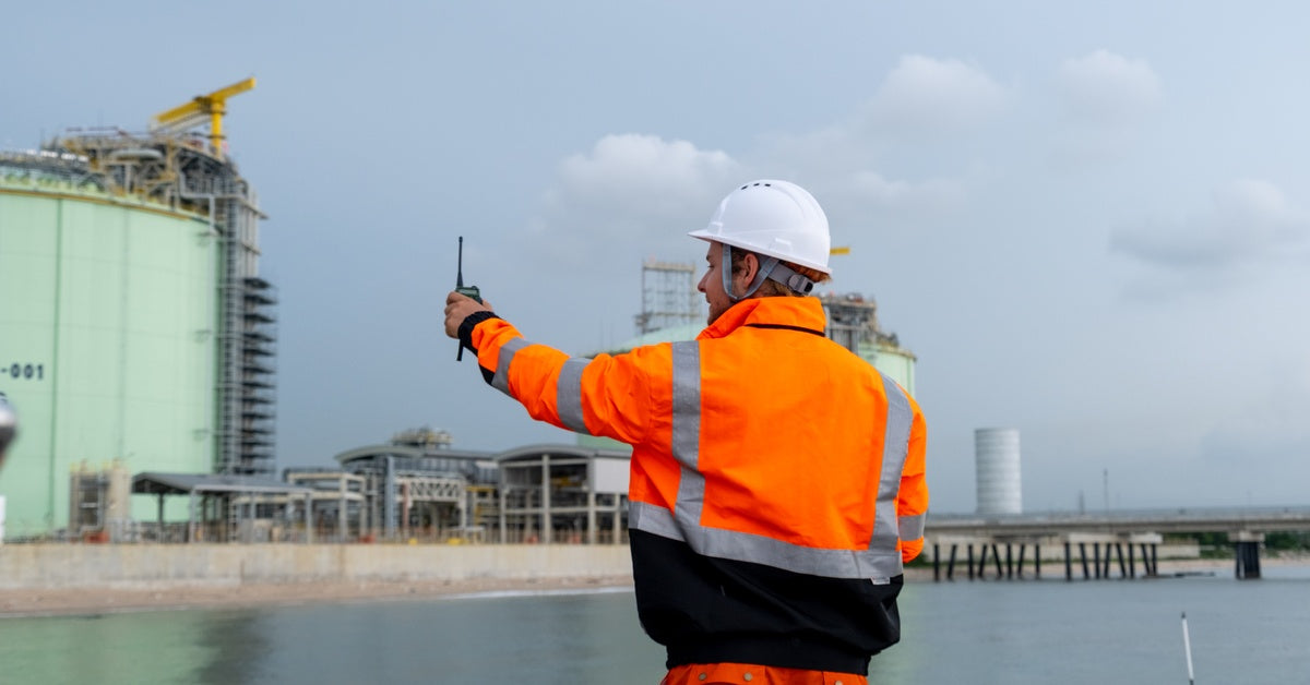 A worker in safety gear standing near an industrial facility and reaching out while communicating with a two-way radio.