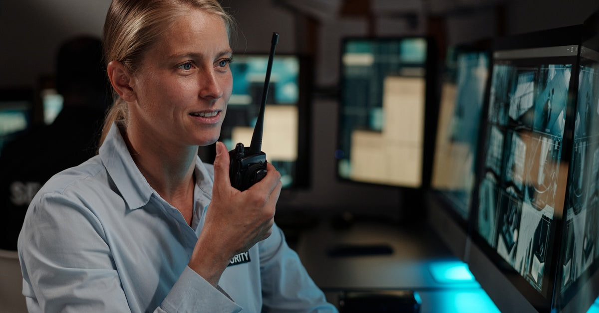 A woman in a blue security shirt speaks into a walkie-talkie while monitoring security footage on desk-mounted screens.