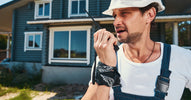 A man stands in the yard of a renovated home while wearing a white hard hat and communicating on a two-way radio.