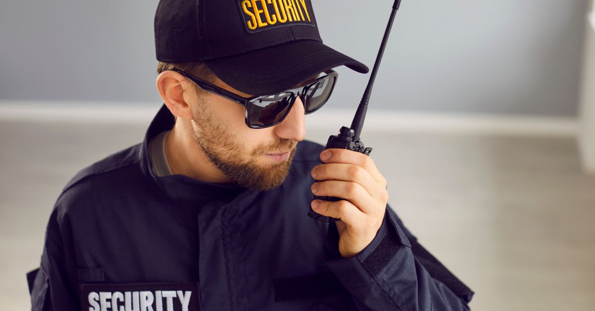A security guard in a blue uniform, hat, and sunglasses stands indoors using a two-way radio to communicate.